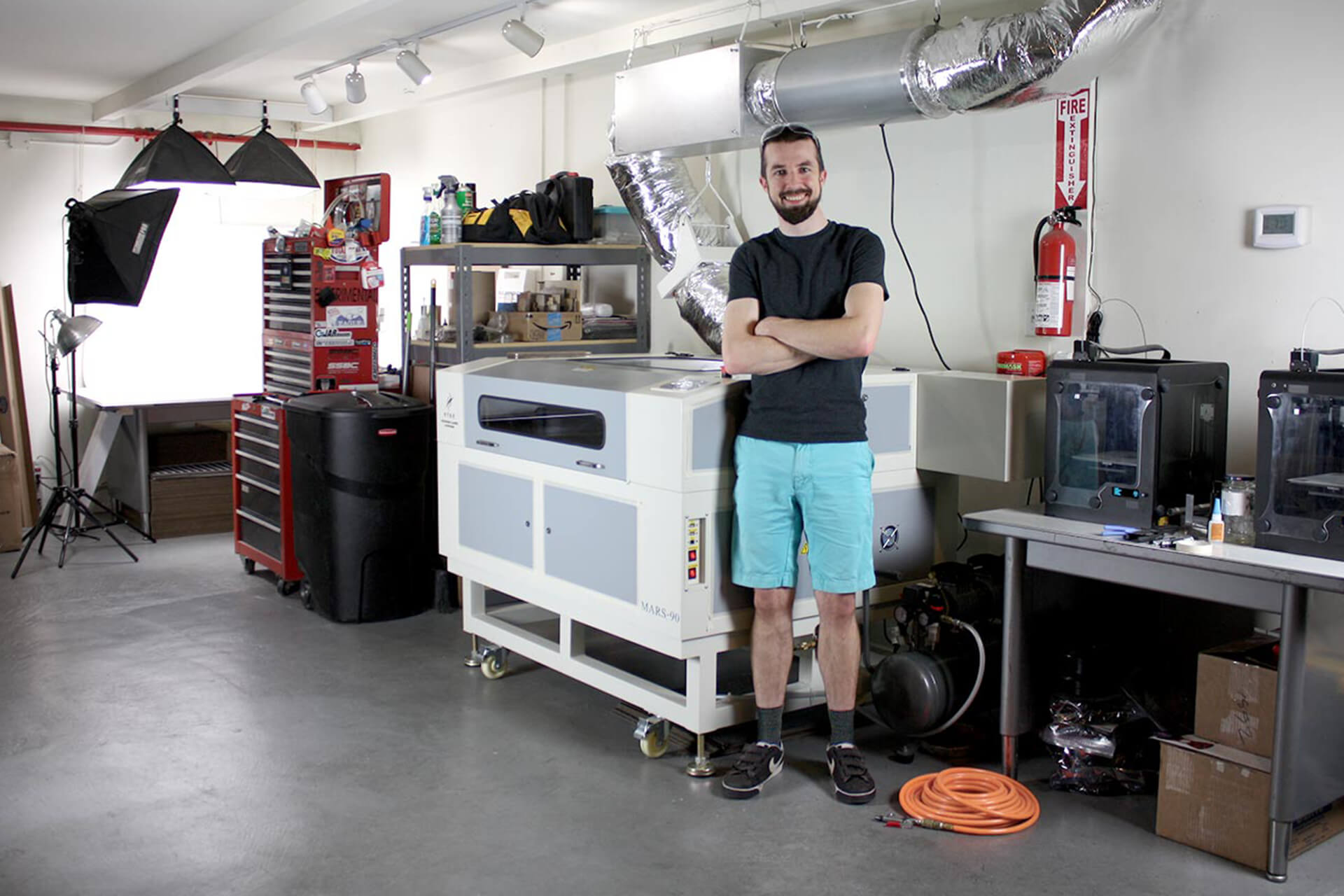 Josh, the owner, smiling and standing in his shop. A laser cutter and 3D printer machines are visible.